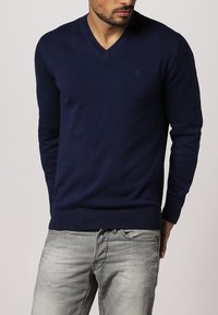 Granatowy sweter typu V-neck, wykonany z miękkiego dzianiny. Posiada żeberkowe mankiety i dół. Dyskretny detal z logo na klatce piersiowej. W połączeniu z jasnoszarymi dżinsami.