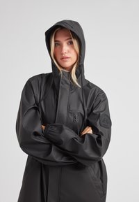 Schwarze wasserdichte Jacke mit Kapuze, ausgestattet mit einem Frontreißverschluss, elastischen Bündchen und einem Logopatch am Ärmel. Glatte, glänzende Textur.