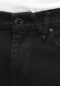 Gros plan sur la ceinture d'un jean en denim noir avec un bouton portant l'inscription « Volcom Brand Jeans » et des passants de ceinture, sur un fond de tissu blanc.