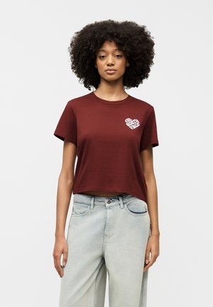 Γυναίκα με σγουρά μαλλιά που φοράει ένα μωβ κόψιμο κορυφής με λευκό λογότυπο "The North Face" και ανοιχτό μπλε ψηλόμεσα τζιν.