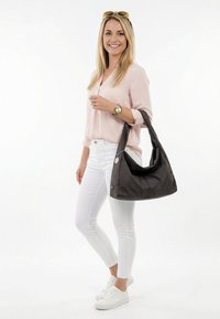 Usmívající se žena s dlouhými blond vlasy, světle růžovou blůzou, bílými kalhotami, bílými teniskami, nesoucí velkou černou kabelku přes rameno a s brýlemi na hlavě.