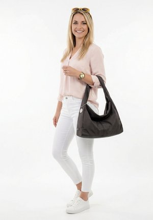 Usmívající se žena s dlouhými blond vlasy, světle růžovou blůzou, bílými kalhotami, bílými teniskami, nesoucí velkou černou kabelku přes rameno a s brýlemi na hlavě.