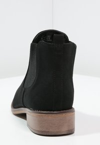 Czarny botek za kostkę o gładkiej, matowej teksturze zamszowej, z elastycznymi bocznymi panelami i jasnobrązowym, warstwowym obcasem. Czysty, minimalistyczny design bez ozdobników.