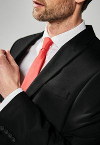 Zwarte suitjack met een gladde textuur, witte shirt en een koraalkleurige das. De das is effen zonder patroon, en de jas heeft één enkele pocket.