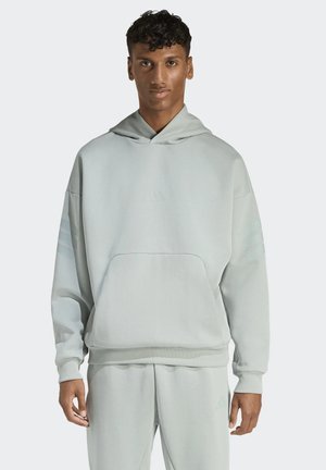 Joven con sudadera con capucha de gran tamaño color gris claro y pantalones a juego, de pie frente a un fondo liso.