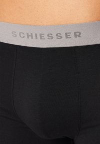 Ropa interior negra ajustada con una cinturilla texturada gris que presenta el nombre de la marca en relieve "SCHIESSER". Tejido suave, costuras mínimas.