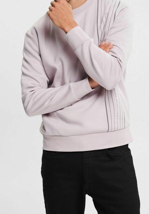 Sudadera de color rosa claro con puños y dobladillo de canalé. Presenta un patrón de líneas diagonales en el lado izquierdo, hecha de tela suave. Pantalones negros.