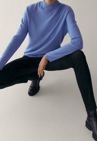 Pull bleu clair à manches longues avec un col montant, associé à un pantalon noir et des bottines, sur fond neutre.