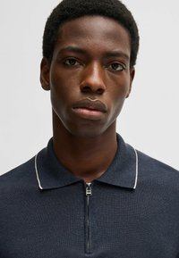 Granatowa koszulka polo wykonana z fakturowanej tkaniny, z zamkiem błyskawicznym przy szyi i białymi akcentami na kołnierzyku. Zbliżenie na odzież.