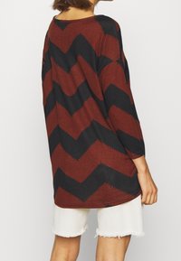 Langärmliges Oberteil mit einem Zickzackmuster in Rost und Schwarz, aus einem weichen Strickmaterial gefertigt. Kombiniert mit weißen, fransigen Shorts.
