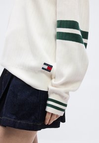 Άσπρο πουλόβερ με πράσινες ρίγες στα μανίκια και στη μέση, μικρό λογότυπο Tommy Jeans κοντά στο τελείωμα, συνδυασμένο με σκουρόχρωμη τζιν φούστα.