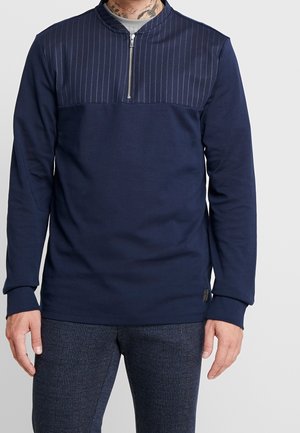 Mężczyzna ubrany w granatową koszulę z długim rękawem z lampasem w prążki i półzamkiem, w połączeniu z teksturowanymi ciemnoniebieskimi spodniami, stojący na tle jednolitego tła.