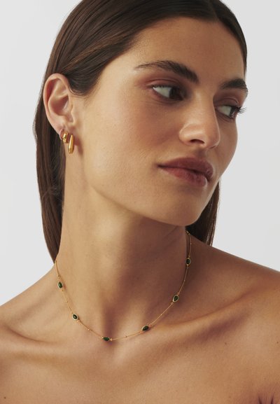 Χρυσό κολιέ με πράσινους λίθους, που διαθέτει μια λεπτή αλυσίδα και οβάλ πολύτιμους λίθους σε τακτά διαστήματα. Απλά χρυσά σκουλαρίκια με κομψό σχέδιο.