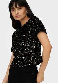 Haut noir à sequins avec manches courtes, présentant une surface texturée et une coupe ample. Porté avec un pantalon noir.