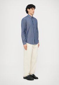 Niebieska koszula chambray zapinana na guziki, z kołnierzykiem, długimi rękawami i subtelną fakturą, noszona z kremowymi spodniami i czarnymi butami.