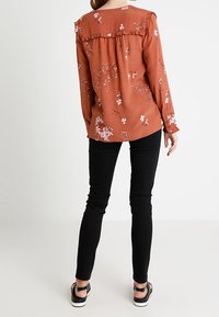 Rostfärgad blus med blommönster, volang vid halsringningen, lång ärm. Tillsammans med svarta tajta byxor och platta svarta sandaler.