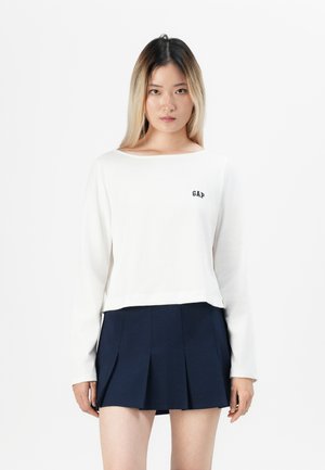 Jeune femme avec des cheveux raides bicolores portant un haut à manches longues blanc avec le logo "GAP" et une jupe plissée bleu marine.
