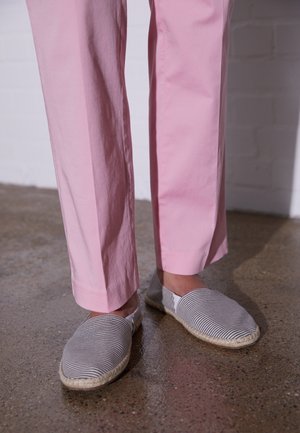 Benen met lichtroze broek en slip-on schoenen met zwart-witte strepen en gevlochten zolen, staand op een gepolijste betonnen vloer.