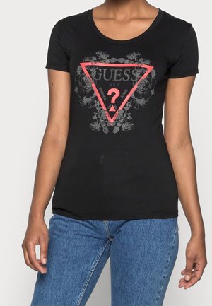 Vrouw die een strakke zwarte T-shirt draagt met een rood driehoek- en bloemenpatroon logo en een blauwe spijkerbroek.