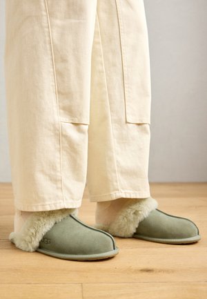 Papuci din suede verde cu căptușeală moale din blană, purtați cu pantaloni de culoare crem, având un design simplu și talpă plată.