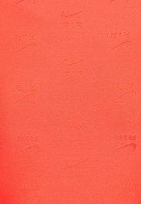 Textil mit einer einheitlichen Korallenfarbe und einem wiederholenden, erhabenen Muster des Nike-Logos sowie dem Wort "AIR" in verschiedenen Ausrichtungen.