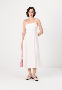 Abito bianco senza maniche con scollatura e orlo a onde, abbinato a una borsa rosa. Il modello indossa scarpe chiare con cinturini.