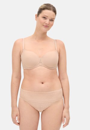 Mujer de piel clara y cabello recogido, de pie, usando un sujetador beige con copas moldeadas y ropa interior a juego con estampado de encaje, frente a un fondo liso.