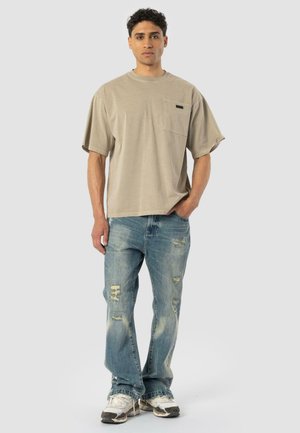 Young man standing, wearing a loose beige t-shirt with a chest pocket, distressed blue jeans, and white sneakers against a plain background.