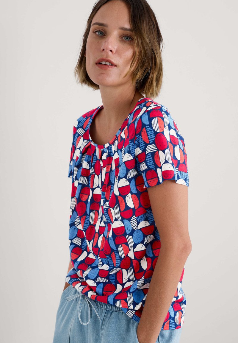 Top de manga corta en algodón azul con patrones abstractos en rojo y blanco. Presenta un escote redondo y un detalle fruncido en el frente. Corte casual.