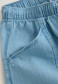 Pantalon en denim bleu clair avec ceinture élastique et couture de poche avant visible, montrant la texture du tissu et les détails des coutures.