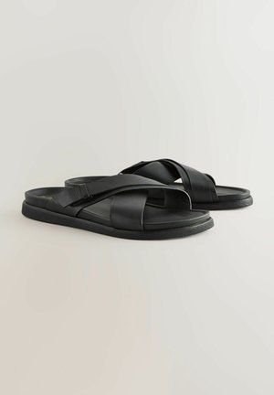 Sandales en cuir noir à glisser avec des brides croisées, semelle plate et finition texturée. Design simple, bout ouvert et style minimaliste.