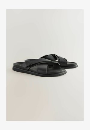 Juodos odinės šlepetės su kryžminėmis juostelėmis, plokščiu padu ir tekstūruotu paviršiumi. Paprastas dizainas, atviras pirštas ir minimalistiškas stilius.