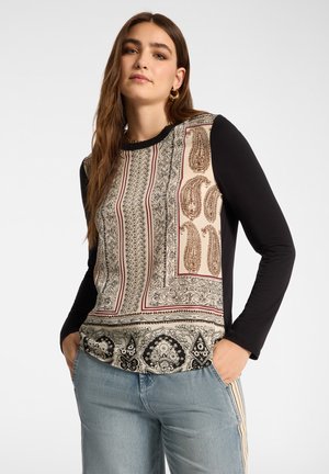 Donna con capelli lunghi castani che indossa una maglia beige e nera a maniche lunghe con motivo e jeans azzurri chiari con le mani nelle tasche.