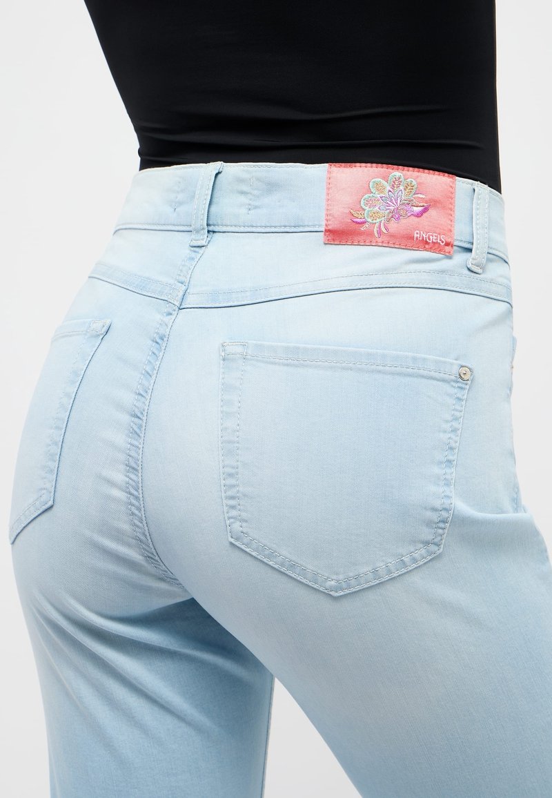 Gros plan d'une personne portant un jean bleu clair avec des poches arrière et une étiquette florale rose indiquant "ANGELS" à la taille.
