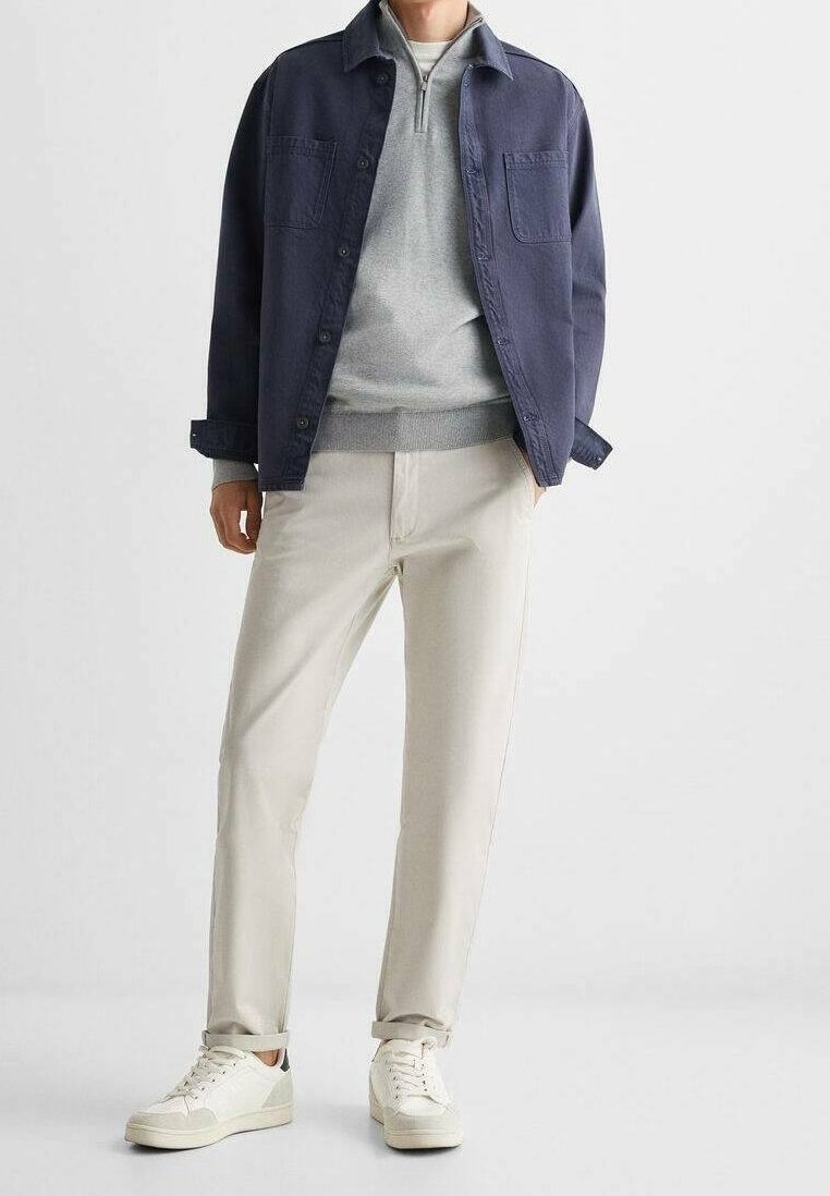 Homme portant une veste bleu marine, un pull gris clair à fermeture éclair au quart, un pantalon beige et des baskets blanches, debout devant un fond blanc.