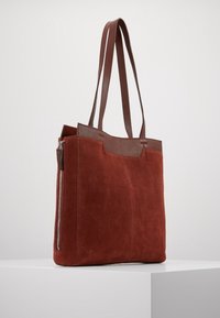 Sac fourre-tout en suede marron avec une finition en cuir lisse sur le dessus, doté de deux longues poignées et d'une poche latérale zippée.