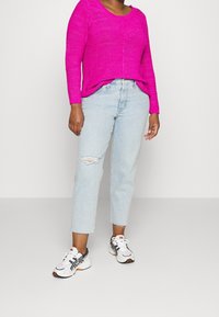 Maglione lavorato a maglia fucsia, jeans chiari strappati e sneakers bianche con dettagli blu navy e arancioni. Vestibilità rilassata, vita a metà.