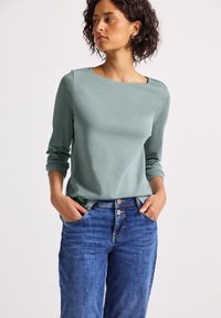 Vrouw met kort, krullend haar die een lichtgroene lange mouwtop en een blauwe high-waisted spijkerbroek draagt, met handen in haar zakken, naar links kijkend.