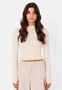 Beige lange mouw top met een hoge hals, voorzien van een voorkeursrits en een cropped ontwerp, gemaakt van een gladde, rekbare stof.
