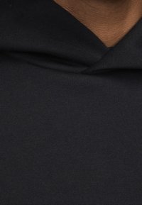 Černá bavlněná mikina s kapucí na stahovací šňůrku a hladkým povrchem. Detailní záběr na látku ukazující stehy a detaily výstřihu.