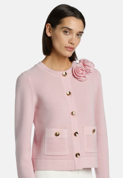 Roza kardigan iz strukturiranega pletenja, opremljen z zlatimi gumbi, dvema sprednjima žepoma in cvetličnim detajlom na rami.