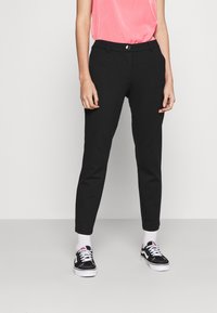 Svarta avsmalnande byxor med en slät yta, med en midja med bältesöglor, framfickor och en silverknapp som detalj. Stylade med svarta sneakers.