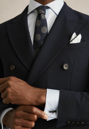 Man adjusting black cufflink on white shirt cuff, wearing dark double-breasted suit with patterned tie and white pocket square.