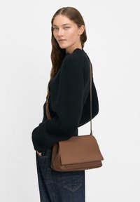 Borsa a spalla in pelle marrone con design a pattina, texture liscia e tracolla sottile regolabile, abbinata a un maglione in lana scuro e jeans.