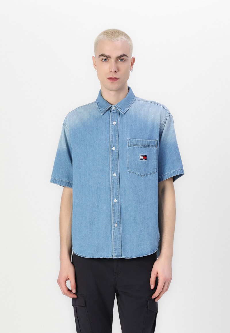 Cămașă din denim cu mânecă scurtă, de culoare albastru deschis, cu închidere prin nasturi, guler ascuțit, un buzunar pe piept și o etichetă logo subtilă în roșu, alb și albastru.