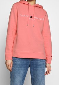 Hoodie rose corail avec un cordon de serrage, logo avant "TOMMY HILFIGER EST. 1985," et manches côtelées. Associé à un denim bleu clair.