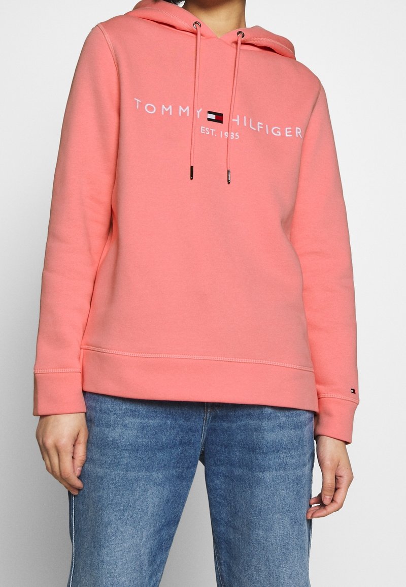 Hoodie rose corail avec un cordon de serrage, logo avant "TOMMY HILFIGER EST. 1985," et manches côtelées. Associé à un denim bleu clair.