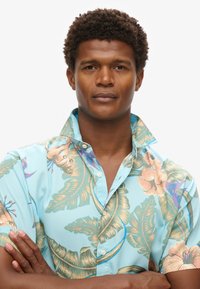 Camicia a maniche corte in tessuto azzurro chiaro con motivi floreali tropicali in rosa e verde, dotata di colletto a punta e chiusura con bottoni.