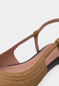 Καφέ υφαντό επίπεδο παπούτσι με λουράκι στο πίσω μέρος και λεπτό λουρί, με ελαφρώς ροζ πάτο, σε θέα από το πλάι σε λευκό φόντο.