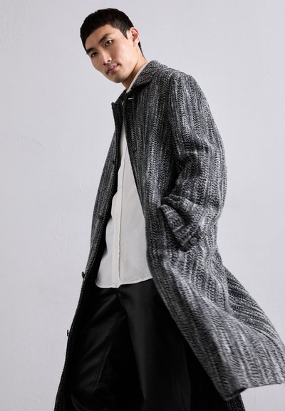 Manteau texturé en tons noir et gris avec un col, des boutons à l'avant et une coupe décontractée, superposé sur une chemise légère et un pantalon noir.